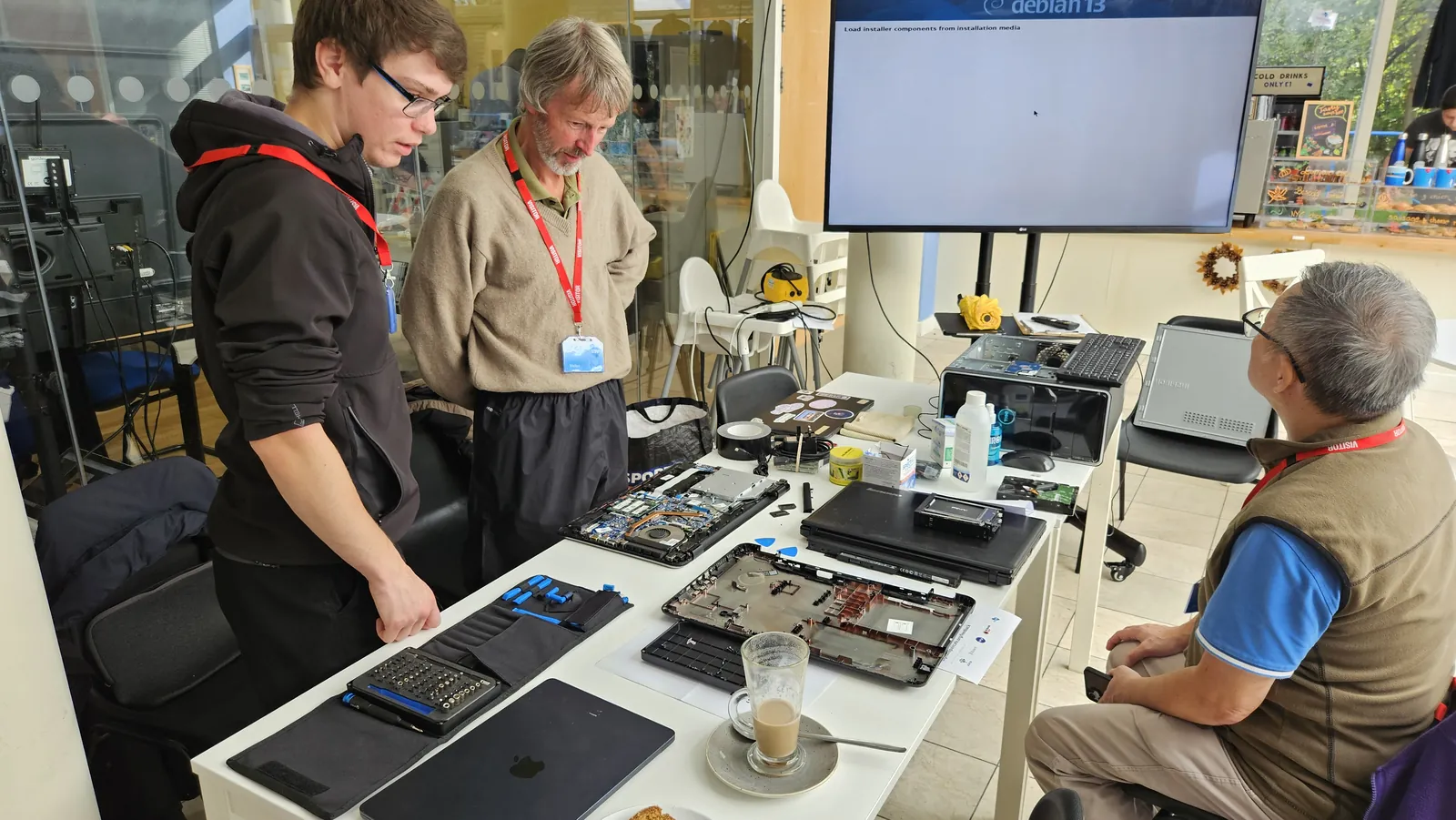 Two people stand at a bench of opened-up laptops while a man in a gilet sits and watches a screen showing the Debian 13 installer.